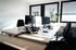 Zwei leere Schreibtische mit Computern, schwarzen Bürostühlen und Pflanzen in einem sonnigen Büro.