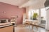 Helles Zimmer mit rosa Wand, Bett, Beistelltisch und Esstisch, Balkontür und Balkon mit Pflanzen.