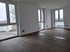 Leerer Raum mit dunklem Holzboden, weißen Wänden und drei großen Fenstern, die auf eine Außenansicht blicken.