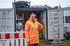Mann in orangefarbener Warnjacke vor offenem Container mit Werkzeugen, gibt Daumen hoch.