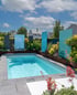 Ein kleiner Pool mit blauer Wasseroberfläche ist von grauen Fliesen und einem Beet mit Pflanzen und blauen Glaswänden umgeben.
