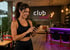 Lachende Frau im Sport-BH blickt auf ihr Smartphone, im Hintergrund ein "Club Vital" Fitnessstudio.