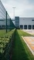 Grüner Zaun, Hecke und Rasen vor einem modernen Lagerhaus mit Laderampen unter blauem Himmel.
