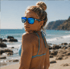 Eine blonde Frau mit Dutt, blauem Bikini und blauen, verspiegelten Sonnenbrillen blickt am Strand über ihre Schulter.