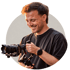 Lächelnder Mann mit dunklem T-Shirt hält eine Kamera auf einem Gimbal-Stabilisator vor hellem Hintergrund.
