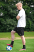 Junger Fußballspieler mit blonden Haaren, weißem T-Shirt und schwarzen Shorts steht mit einem Fuß auf einem Fußball.