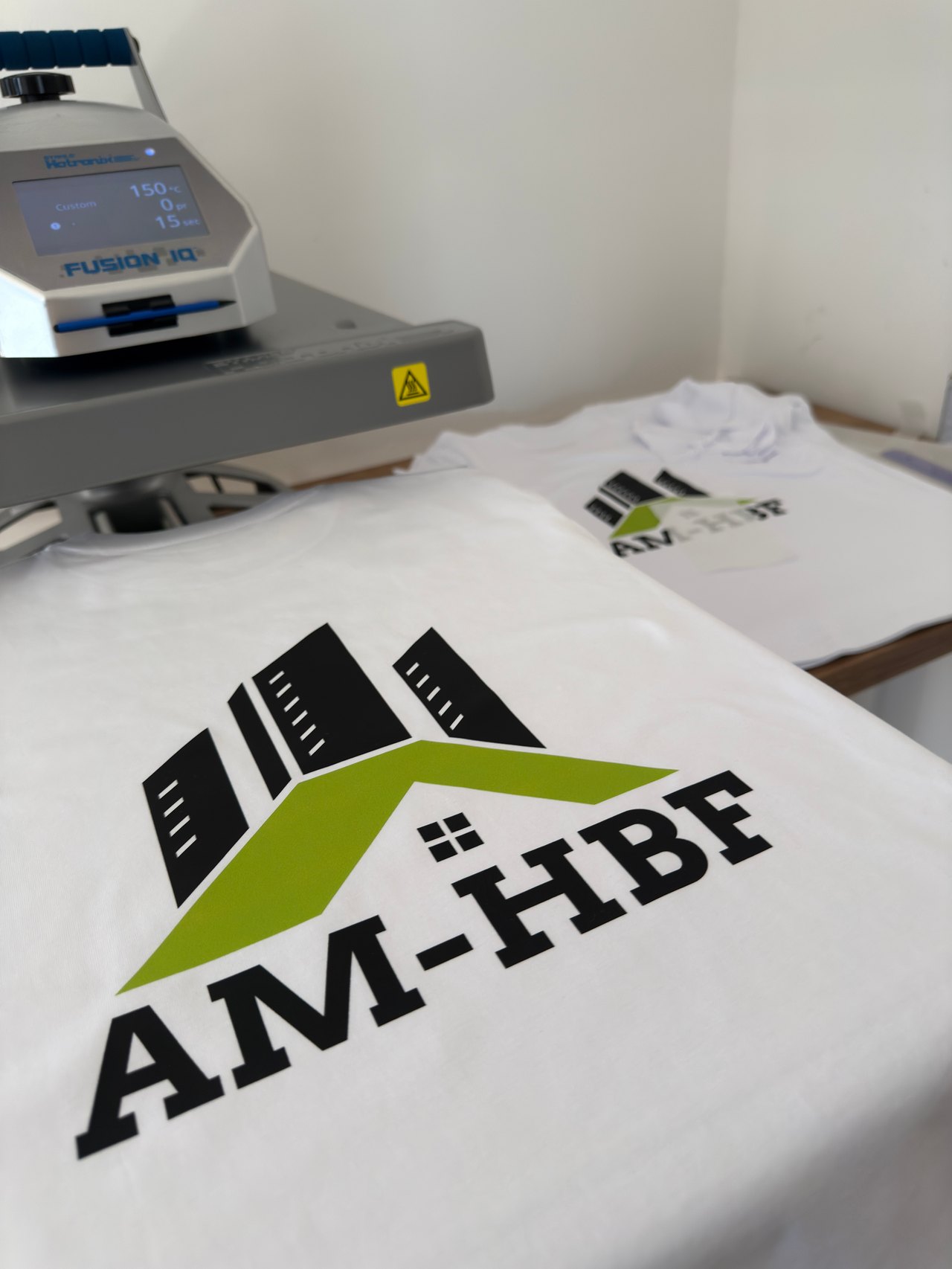 Weißes T-Shirt mit AM-HBF Logo neben der Transferpresse