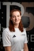 Eine lächelnde Frau mit rötlichem Haar trägt ein weißes Adidas-Sportshirt mit dem Logo von Therapier und Pier5 
