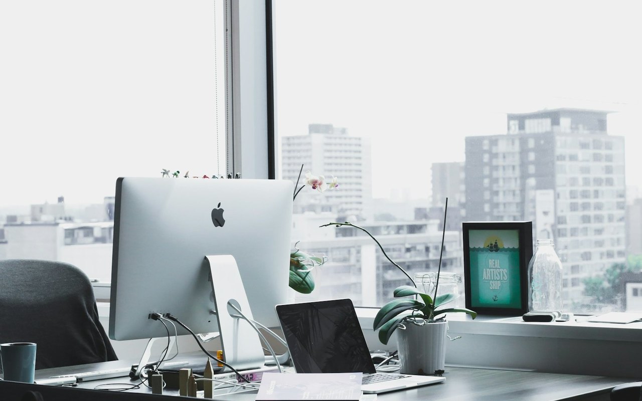 Silberner iMac auf einem Schreibtisch vor einem Fenster mit Stadtblick, daneben ein Laptop