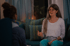 Eine Frau mit Brille und langen Haaren gestikuliert sprechend zu einer anderen Person auf einem Sofa.