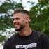 Lächelnder Mann mit Bart und kurzem Haarschnitt trägt ein schwarzes Sweatshirt mit „STRADA ATHLETICS“ Schriftzug.