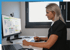 Blonde Frau sitzt am Schreibtisch, arbeitet mit Grafiktablett und Stift vor einem Computermonitor mit Tabellen.