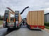 Zwei Lastwagen mit Holzmodulen auf der Ladefläche und einem Kran, der ein Modul auf den rechten LKW hebt.