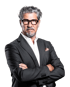 Mann mit grauen Locken, Bart, Brille, weißem Hemd und dunklem Anzug mit verschränkten Armen.