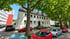 Ein weißes Mehrfamilienhaus mit dunklem Dach hinter grünen Bäumen und geparkten Autos an einem sonnigen Tag.