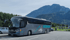 Grauer Reisebus "HÜBNER" parkt auf einem asphaltierten Parkplatz vor hohen, bewaldeten Bergen.