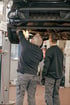 Zwei Männer in Arbeitskleidung inspizieren die Unterseite eines angehobenen Autos in einer Werkstatt.