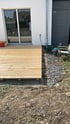 Holzterrasse im Bau vor einer gläsernen Terrassentür an einer weißen Hauswand.