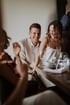 A laughing man in a light suit and a woman in a white dress sit at a table with wine glasses.
