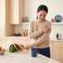 A woman in a kitchen winces while holding her shoulder next to a bag of groceries.