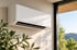 Weiße, moderne Split-Klimaanlage an einer hellen Wand neben einem Fenster mit Blick ins Grüne.