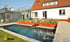 Moderner Garten mit rechteckigem Schwimmbecken, Holzterrasse, Pflanzenbeeten und weißen Wohngebäuden im Hintergrund.