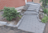 Ein neuer, grauer Steinweg führt zu einer Treppe mit vier Stufen und einer Plattform, daneben ein Busch.