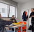 Drei Männer sitzen oder stehen in einem Büro; einer erklärt etwas, während die anderen zuhören.