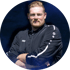 Porträt eines Mannes mit rötlichen Haaren und Bart im schwarzen Trainingsanzug mit verschränkten Armen vor dunklem Hintergrund.