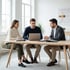 Drei Personen in Business-Kleidung sitzen an einem Holztisch mit Laptop und Aktenordnern in einem hellen Büro.