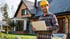 Lächelnder Mann im gelben Schutzhelm und kariertem Hemd hält Laptop vor einem Haus mit Solarmodulen.