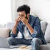 Man in denim shirt with glasses rubbing eyes at laptop on couch, showing signs of chronic fatigue