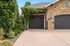 A stone house with two dark gray garage doors, a concrete driveway, and green landscaping with orange flowers.