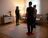 Drei Personen stehen auf farbigen Matten in einem Wohnzimmer, zwei weitere Matten liegen leer.
