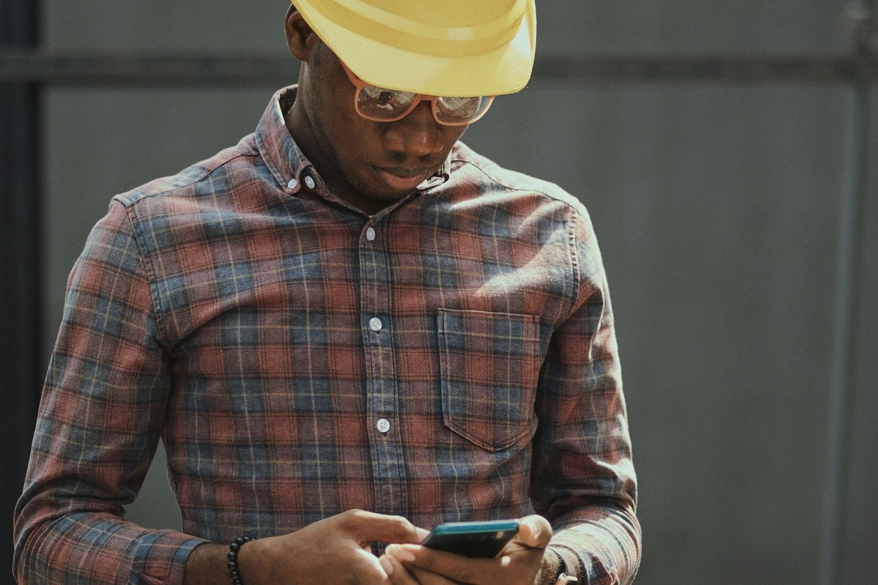 Handwerker mit gelbem Helm und kariertem Hemd tippt auf seinem Smartphone