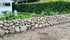 Eine Trockenmauer aus runden Steinen grenzt einen Garten mit Hecke und weißem Haus ab.