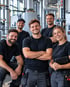 Fünf lächelnde Handwerker in schwarzer Arbeitskleidung posieren vor industriellen Rohrleitungen und Anlagen.