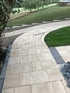 Ein geschwungener Gartenweg aus hellen Pflastersteinen führt an einer Rasenfläche und Bäumen vorbei hangabwärts.