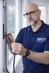 Ein Mann in blauem Hemd mit "BUFU Fenstertechnik München"-Logo montiert eine schwarze Dichtung an einem Fensterrahmen.