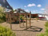 Spielplatz mit Holzklettergerüst und Schaukeln auf Rindenmulch vor Häusern und blauem Himmel.