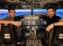 Zwei Männer sitzen im Cockpit eines Flugsimulators vor einem Bildschirm, der eine Stadtansicht zeigt.