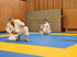Zwei Männer in weißen Kimonos auf einer blauen Matte in einem Trainingsraum mit Holzpaneelen.