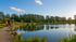 Ein ruhiger See mit grünen Bäumen und einem kleinen Holzhaus am Ufer unter blauem Himmel mit weißen Wolken.