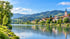 Stadt am Flussufer mit Kirchturm vor bewaldeten Bergen und blauem Himmel bei Sonnenschein.