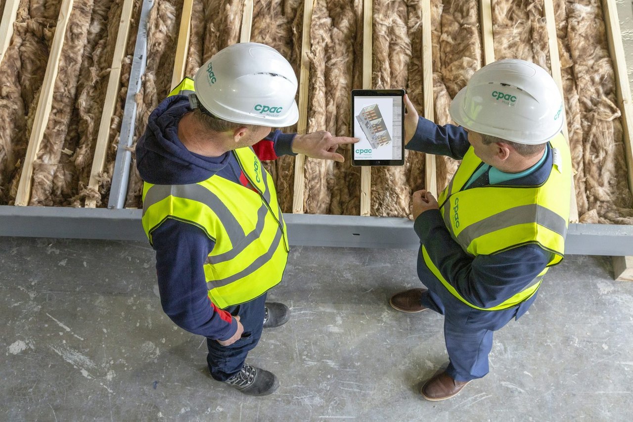Zwei Bauarbeiter mit Helm und Warnweste schauen gemeinsam auf ein Tablet vor einer gedämmten Holzwand