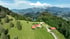 Alpenlandschaft mit grünen Wiesen, bewaldeten Hügeln, einem See und einem Gebäudeensemble.