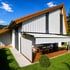 Modernes Haus mit Holzboden-Terrasse, weißer Markise und vertikalem Sichtschutz an sonnigem Tag.