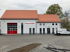 Graues Feuerwehrhaus mit rotem Tor und "Feuerwehr" Schriftzug; weißer Lieferwagen parkt davor.