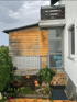 Haus mit Holzverkleidung, Ziegelmauer, Balkon, einer Tür und einem kleinen Gartenbeet.