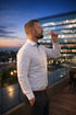 Ein Mann mit Bart in weißem Hemd trinkt Sekt auf einer Dachterrasse bei Sonnenuntergang vor einer Stadtkulisse.